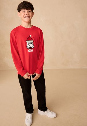 Besijuokiantis berniukas raudonu „Star Wars“ megztiniu su Kalėdų senelio kepurės Stormtrooperio dizainu, juodomis kelnėmis ir baltais sportbačiais, stovintis prie smėlio spalvos fono.