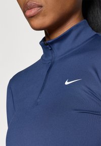 Tumši zils sporta tops ar ceturtdaļas rāvējslēdzēju un mazu baltu Nike logo uz krūtīm. Gluda, elastīga auduma materiāls ar pieguļošu formu.