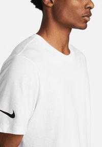 Weißes Baumwoll-T-Shirt mit kurzen Ärmeln, rundem Halsausschnitt und einem schwarzen Nike-Logo auf dem linken Ärmel. Glatte Textur und klassischer Schnitt.