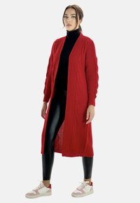 Röd stickad kofta med fläta, öppen framsida och långa ärmar, kombinerad med svarta leggings och vita sneakers med rosa accenter.