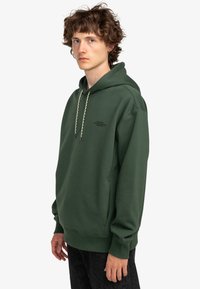 Hoodie en coton vert avec un cordon de serrage, des poignets et un ourlet côtelés. Présente un petit logo noir sur la poitrine gauche. Le modèle porte un jean foncé.