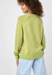 Grüner gestrickter Pullover mit einem gerippten Saum und Bündchen, ausgestattet mit einem Raglan-Ärmel Design und weichem Material. Über einer hellblauen Jeans getragen.