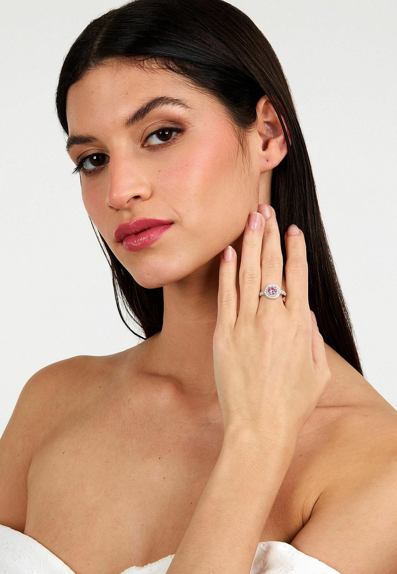 Anello d'argento con una gemma rotonda centrale rosa, circondata da piccole pietre chiare, montato su una fascia testurizzata. Il modello lo mostra sulla sua mano.