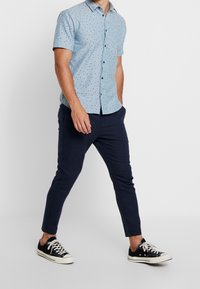 Man som går iklädd en ljusblå kortärmad skjorta med små svarta mönster, marinblå slim-fit byxor och svarta låga sneakers.