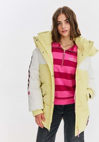 Padded jacket v žltej a bielej farbe s vysokým golierom, predným zipsom, bočnými vreckami a čiernymi akcentmi na rukávoch. Pod ním pruhovaný ružový pulóver.