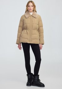 Beżowa kurtka puchowa z pluszowym kołnierzem, zaciśniętą w pasie oraz pikowanym wykończeniem. W zestawieniu z czarnymi legginsami i czarnymi botkami z futerkiem.