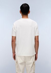 Mann trägt ein schlichtes weißes Kurzarm-T-Shirt und cremefarbene Cargo-Hose und steht mit dem Rücken zur Kamera vor einem hellgrauen Hintergrund.