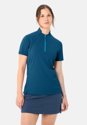 Frau mit braunen Haaren, die ein blaues kurzärmeliges, mit Reißverschluss versehenes Oberteil und einen dunkelblauen Rock trägt, steht vor einem einfarbigen hellgrauen Hintergrund.