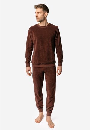 Mann steht barfuß, trägt ein passendes braunes Langarmshirt und Hose aus weichem Stoff und schaut nach links.