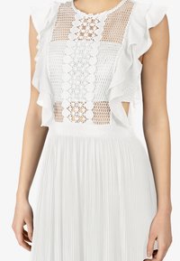 Biała sukienka z fakturowaną górą ozdobioną koronką i wycięciami, z falbanianymi rękawami oraz plisowaną spódnicą. Wykonana z lekkiego materiału.
