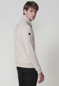 Giovane uomo in giacca zip-up beige a trama con colletto alto e pantaloni neri, girato leggermente di lato, che guarda oltre la spalla.