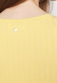 Gros plan sur le haut du dos d'une femme portant un haut jaune côtelé avec un petit pendentif en métal en forme de cœur près de l'encolure.