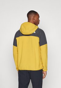 Kollane ja hall fliisist kapuutsiga pusa, millel on kontrastne õlavarre disain, sile tekstuur ja The North Face logo seljal.