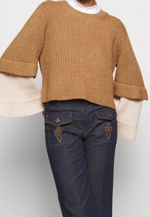 Žena v vrstevnatém béžovém a krémovém svetru s rukávy do zvonu a vysokým krajkovým límcem, doplněném tmavě modrými džínami s vyšívanými kapsami s klopou.