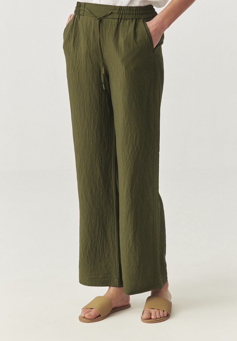 Olijfgroene, wijde broek met een elastische tailleband, gestructureerde stof en zijzakken. Gecombineerd met tankleurige slidesandalen.