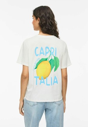 Kvinde med mørkt hår iført hvid T-shirt med stor gul citron, grønne blade og blå tekst "CAPRI ITALIA" på ryggen, kombineret med lyseblå jeans.