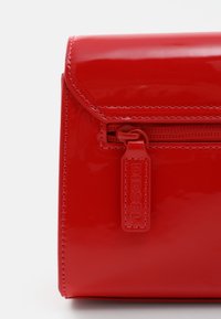 Sac en simili cuir rouge verni avec une fermeture à rabat, doté d'une poche zippée à l'avant et d'une tirette de fermeture éclair marquée. Texture lisse et brillante avec des coutures soignées.