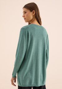 Žena s dlouhými hnědými vlasy, nosící volný tyrkysový svetr s dlouhým rukávem, stojící bokem proti jednolitému béžovému pozadí.