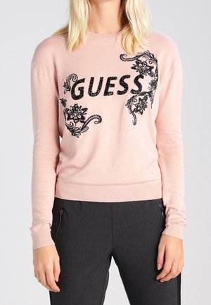 Jasnoróżowy sweter z czarnym kwiatowym haftem i logo "GUESS" na piersi, noszony z czarnymi spodniami z kieszeniami na suwak.