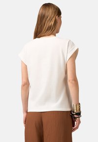 Weißes Baumwoll-T-Shirt mit umgeschlagenen kurzen Ärmeln, das eine lockere Passform und eine glatte Textur aufweist, kombiniert mit braunen plissierten Hosen.
