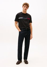 Mladi moški z kodrastimi lasmi, oblečen v črno majico Tommy Hilfiger, temne kavbojke in rjave loaferje, stoji z eno roko v žepu.