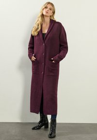 Lång lila cardigan med knappfäste, två framfickor och ribbade ärmslut, matchad med svarta ankelstövlar och en figurnära topp.