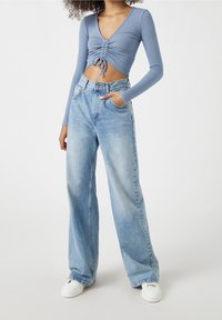 Helles blaues, geripptes langärmliges Oberteil mit gerafftem Vorderteil, kombiniert mit hoch taillierten, weit geschnittenen, ausgebleichten blauen Jeans und weißen Sneakers.