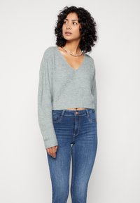 Suéter corto gris claro con cuello en V, textura acanalada y mangas largas, combinado con jeans ajustados azules.