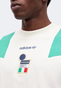 Άνδρας που φοράει λευκό πουκάμισο Adidas με πράσινες λεπτομέρειες στους ώμους και το λογότυπο της ιταλικής ποδοσφαιρικής ομοσπονδίας με τέσσερα αστέρια και σημαία.