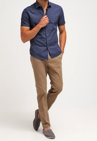 Camicia navy a maniche corte con pois bianchi, pantaloni khaki e scarpe slip-on grigie. La camicia presenta un design con bottoni e una vestibilità su misura.