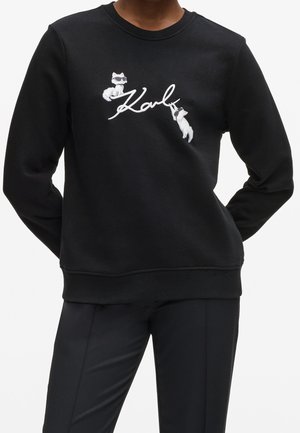 Czarna bluza z dwiema białymi, kreskówkowymi kotami w okularach przeciwsłonecznych wokół kursywnego słowa „Karl" na piersi, noszona przez stojącą osobę.