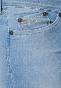 Light blue denim jeans featuring a structured design, front pocket with stitching, and branded tag in gold thread. Black hardware accent.