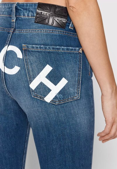 Particolare di jeans in denim blu con lettere bianche dipinte "C" e "H" vicino alla tasca posteriore e una etichetta in pelle nera sopra.