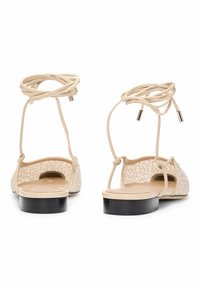 Beżowe płaskie sandały z fakturowaną cholewką i wiązaniami na kostce. Posiadają ozdoby po bokach oraz czarną podeszwę. Otwarty tył.