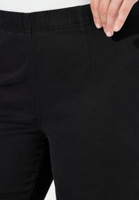 Czarne spodnie elastyczne z elastycznym paskiem w talii, gładką teksturą i subtelnymi detalami szwów. Tkanina ma delikatny połysk.