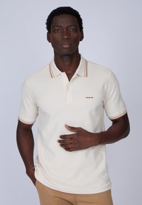 Polo beige avec col et manches rayés marron, fabriqué en tissu texturé. Présente un petit motif brodé sur la poitrine.