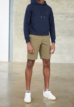 Homme portant un sweat à capuche bleu marine, un short kaki et des baskets blanches, debout sur un sol en béton poli devant un mur en brique beige.