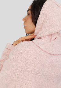 Vrouw in profiel die haar nek aanraakt, gekleed in een lichtroze gemêleerde fleece hoodie met de capuchon omhoog voor een eenvoudige grijze achtergrond.