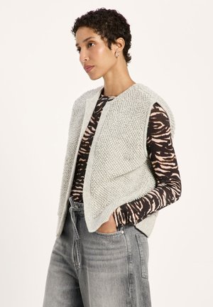 Woman with short curly hair wearing silver hoop earrings, a gray textured knit vest, animal print long-sleeve top, and gray high-waisted jeans with hands in pockets.