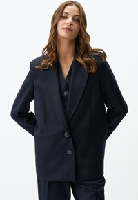 Blazer navy con design doppiopetto, rever a punta e due tasche frontali. Realizzato in tessuto liscio, presenta tre bottoni grandi.