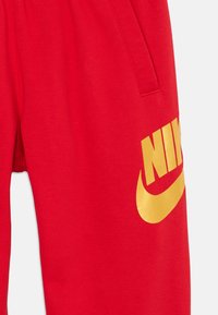 Červené teplákové nohavice vyrobené z mäkkej tkaniny, s veľkým zlatým logom Nike na ľavej nohe a bočnými vreckami. Elastický pás s šnúrkou.