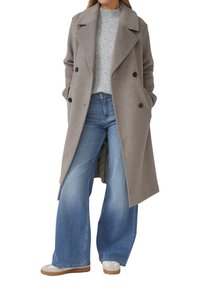 Vrouw in een lange grijze dubbele jas, lichtgrijze gebreide trui, wijde blauwe jeans en witte sneakers. Handen in de zakken van de jas.