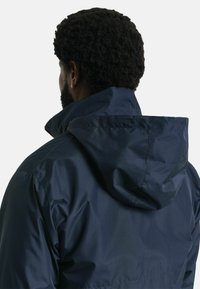 Chaqueta impermeable de color azul marino con un alto collar y capucha ajustable; textura suave con costuras visibles y un corte relajado.