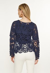 Top croșetat de culoare albastru închis cu mâneci lungi, având modele intricate și un țesut deschis. Designul include un tiv zimțat și un decolteu rotund.