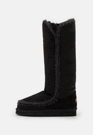 Bottes noires en daim montantes avec des coutures décoratives le long des coutures, une semelle plate et un design à bout rond.