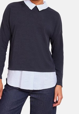 Personne portant un pull à manches longues bleu marine superposé à une chemise blanche à col avec de fines rayures bleues et un jean bleu foncé.