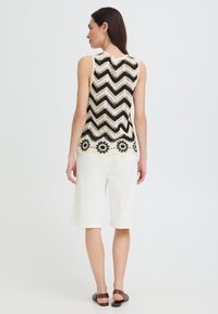 Femme portant un haut sans manches en crochet à motif zigzag noir et blanc avec détail floral à l'ourlet, short blanc longueur genoux et sandales marron.