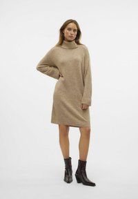 Abito maglione beige con collo alto, vestibilità ampia e maniche lunghe. Indossato con stivaletti neri con tacco. Texture liscia e design casual.
