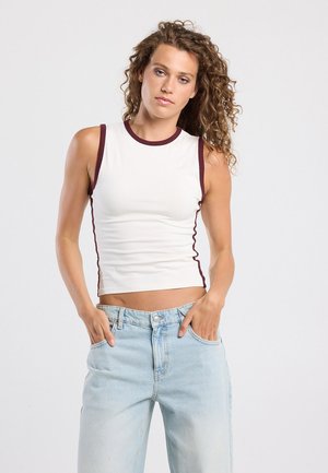 Witte mouwloze top met bordeauxrode rand en geschulpte zijaccenten, gecombineerd met lichtblauwe high-waisted jeans. Gladde stof, fitted ontwerp.