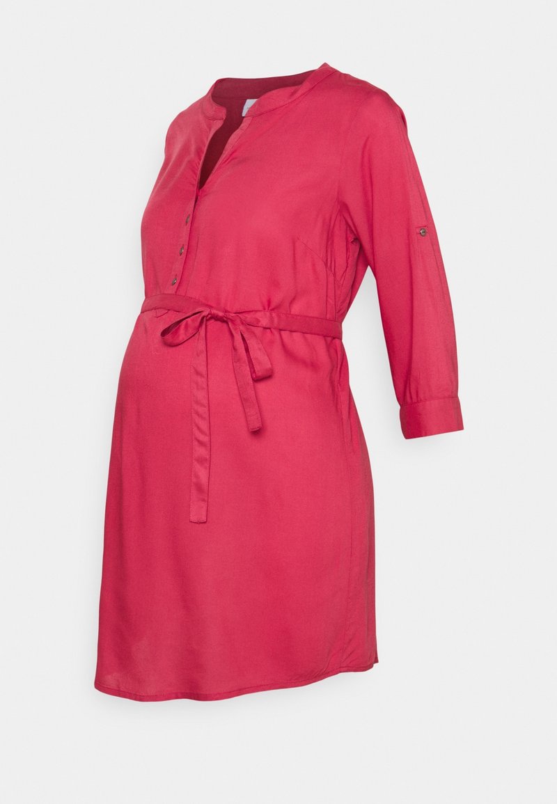 Robe de maternité rouge à manches trois-quarts, col boutonné et ceinture nouée à la taille sur un fond uni.